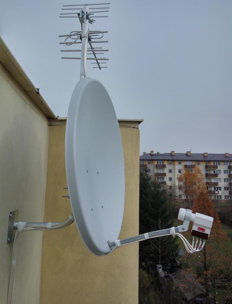 Antena satelitarna i antena naziemna zamontowane na ścianie budynku, z widocznym zewnętrznym dekoderem i okablowaniem zabezpieczonym taśmą, na tle bloku mieszkalnego i drzew bez liści.