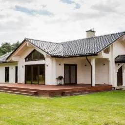 Nowoczesny dom z bali, biała elewacja, ciemna dachówka, drewniany taras, grill na zewnątrz. Widok z trawnika na tle drzew.