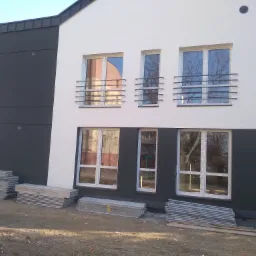 Nowoczesna elewacja dwukondygnacyjnego budynku w Dąbrowie Tarnowskiej, z czarno-białymi panelami i metalowymi balustradami okiennymi, z przygotowanymi rusztowaniami u podstawy.