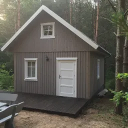 Mały, szary domek z drewnianą elewacją i białymi oknami w leśnym otoczeniu w Warszawie, z widocznym tarasem i fragmentem ławki.