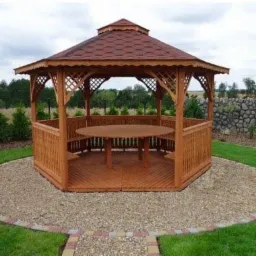 Ośmiokątna altana ogrodowa z drewna, z brązowym dachem z gontów bitumicznych, okrągłym stołem wewnątrz i wbudowanymi ławkami, otoczona trawnikiem i ścieżką z kamyków.
