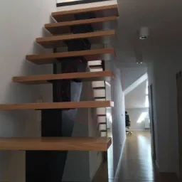 Nowoczesne schody drewniane z czarnymi wspornikami zamontowane przy białej ścianie, widok z dołu na górę, minimalistyczny design.