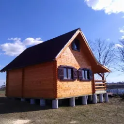 Gotowy drewniany domek letniskowy z brązowym dachem i okiennicami, posadowiony na betonowych słupach, z widokiem na jezioro w tle.