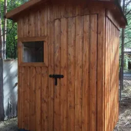 Mała, drewniana budowla z oknem i zaryglowanymi drzwiami, prawdopodobnie szopa lub domek narzędziowy, wykonana z pionowo ułożonych desek w odcieniach brązu, z widocznym zarysem drzew w tle.
