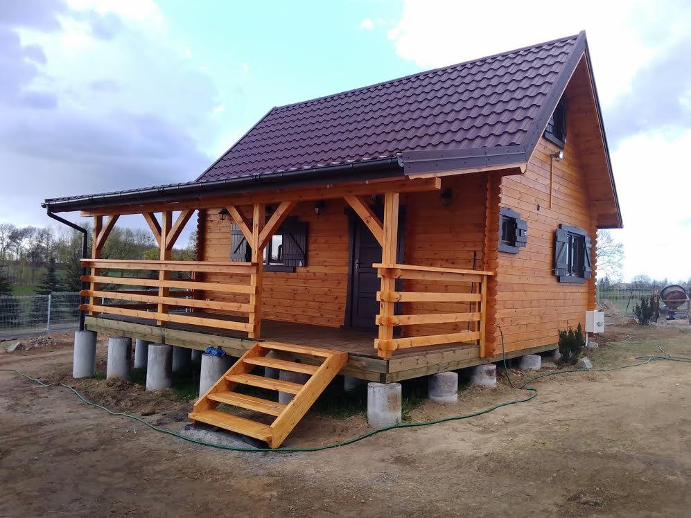 Nowoczesny, drewniany dom z brązowym dachem, podniesiony na betonowych słupach, z drewnianymi schodami i zadaszonym tarasem, widziany z zewnątrz w pochmurny dzień.