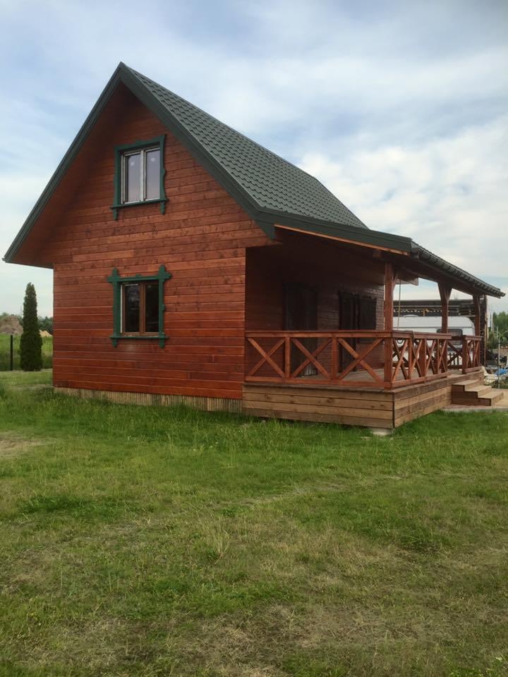 Gotowy, niewielki, parterowy budynek z drewna, z zadaszonym tarasem, pomalowany na brązowo, z zielonymi obramowaniami okien i ciemnozielonym dachem z blachy, otoczony trawnikiem.