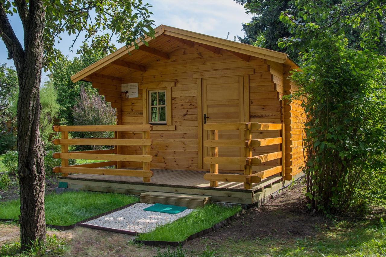 Mały, parterowy domek z drewna, z zadaszonym gankiem, otoczony zielenią. Wejście wyłożone białym żwirem i zielonym dywanikiem.