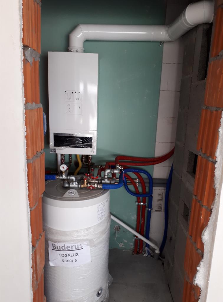 Biały wiszący piec gazowy Buderus z podłączonym zasobnikiem ciepłej wody Logalux S100/5, rurami w kolorze niebieskim i czerwonym oraz białym kominem w surowym wnętrzu z ceglanymi ścianami.