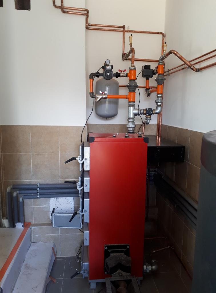 Czerwony piec CO z podłączoną instalacją miedzianą i szarym zbiornikiem wyrównawczym w pomieszczeniu z beżowymi płytkami.