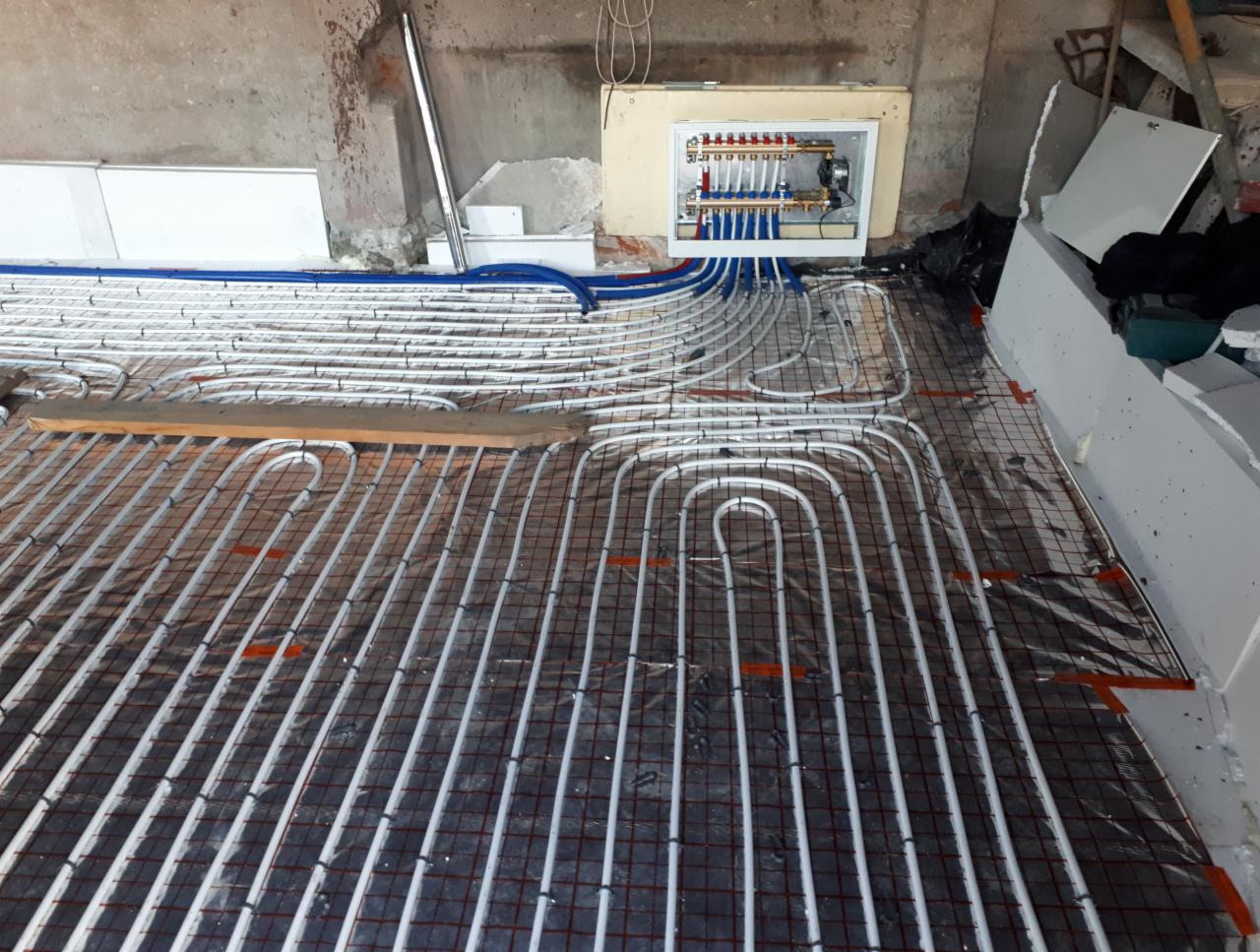 Instalacja ogrzewania podłogowego w trakcie montażu: białe rurki ułożone w pętle na metalowej siatce z folią izolacyjną, rozdzielacz z zaworami zamontowany na ścianie w tle.