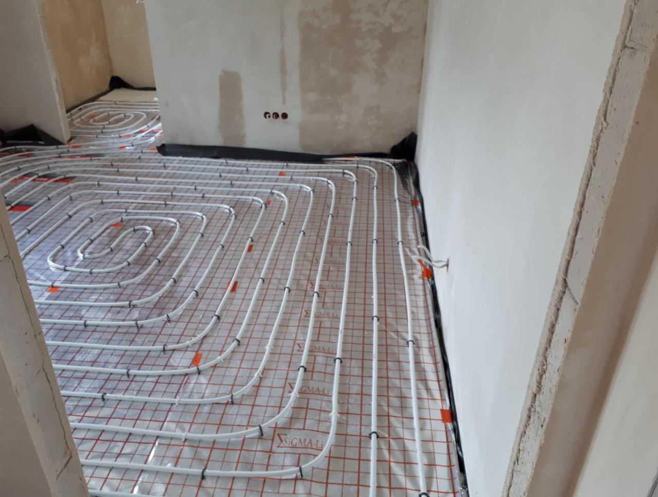 Rozłożona instalacja ogrzewania podłogowego z białych rurek na folii z czerwoną siatką w surowym wnętrzu, widoczne niedokończone ściany i wystające przewody.