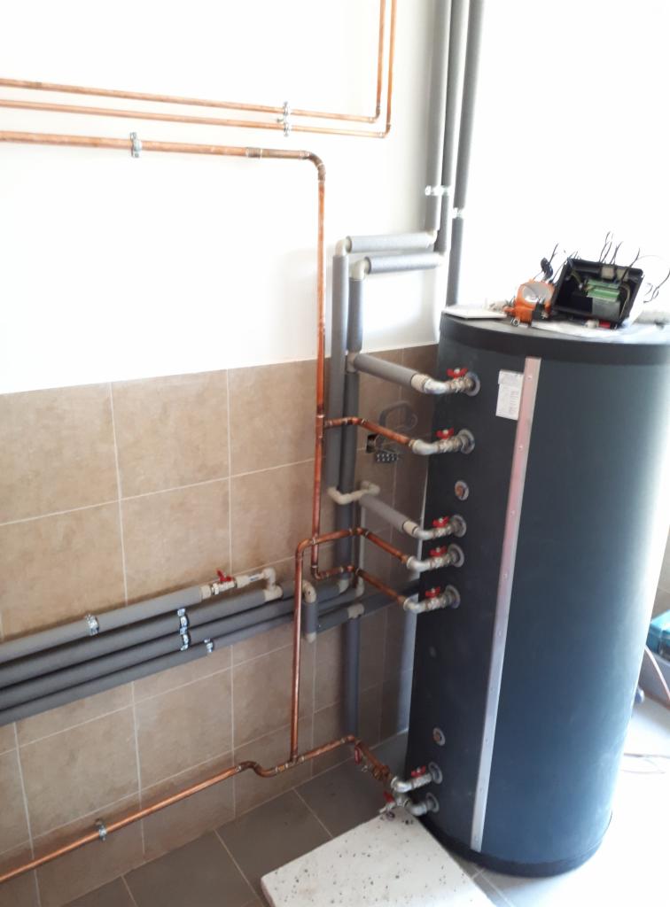 Instalacja centralnego ogrzewania z miedzianymi rurami, szarymi rurami z izolacją i czarnym zbiornikiem akumulacyjnym, widoczne zawory z czerwonymi pokrętłami, zamontowana na ścianie wyłożonej...