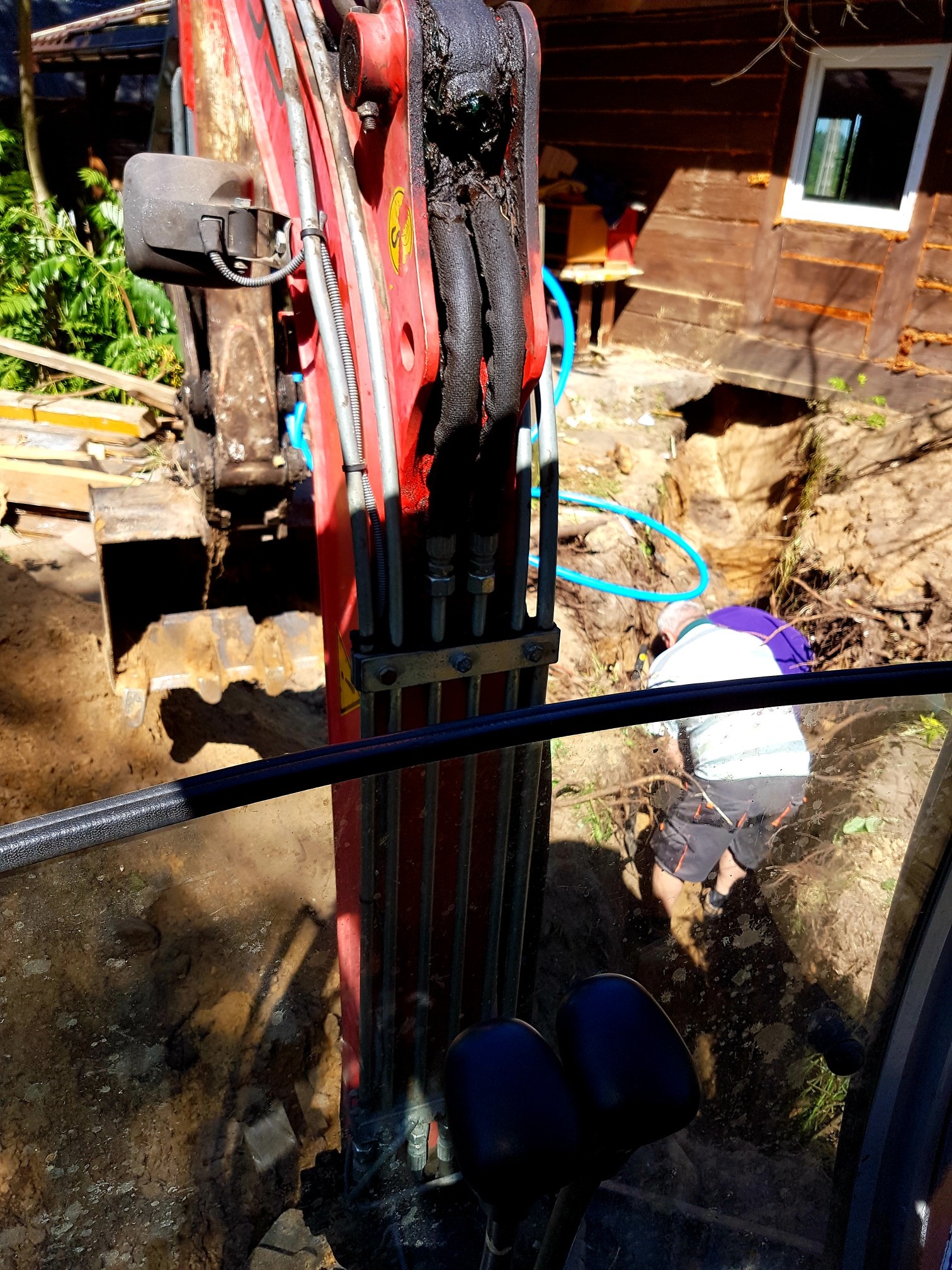 Widok z kabiny koparki na wykop, gdzie mężczyzna pracuje przy układaniu niebieskiego węża w ziemi, obok widoczny fragment drewnianego budynku.