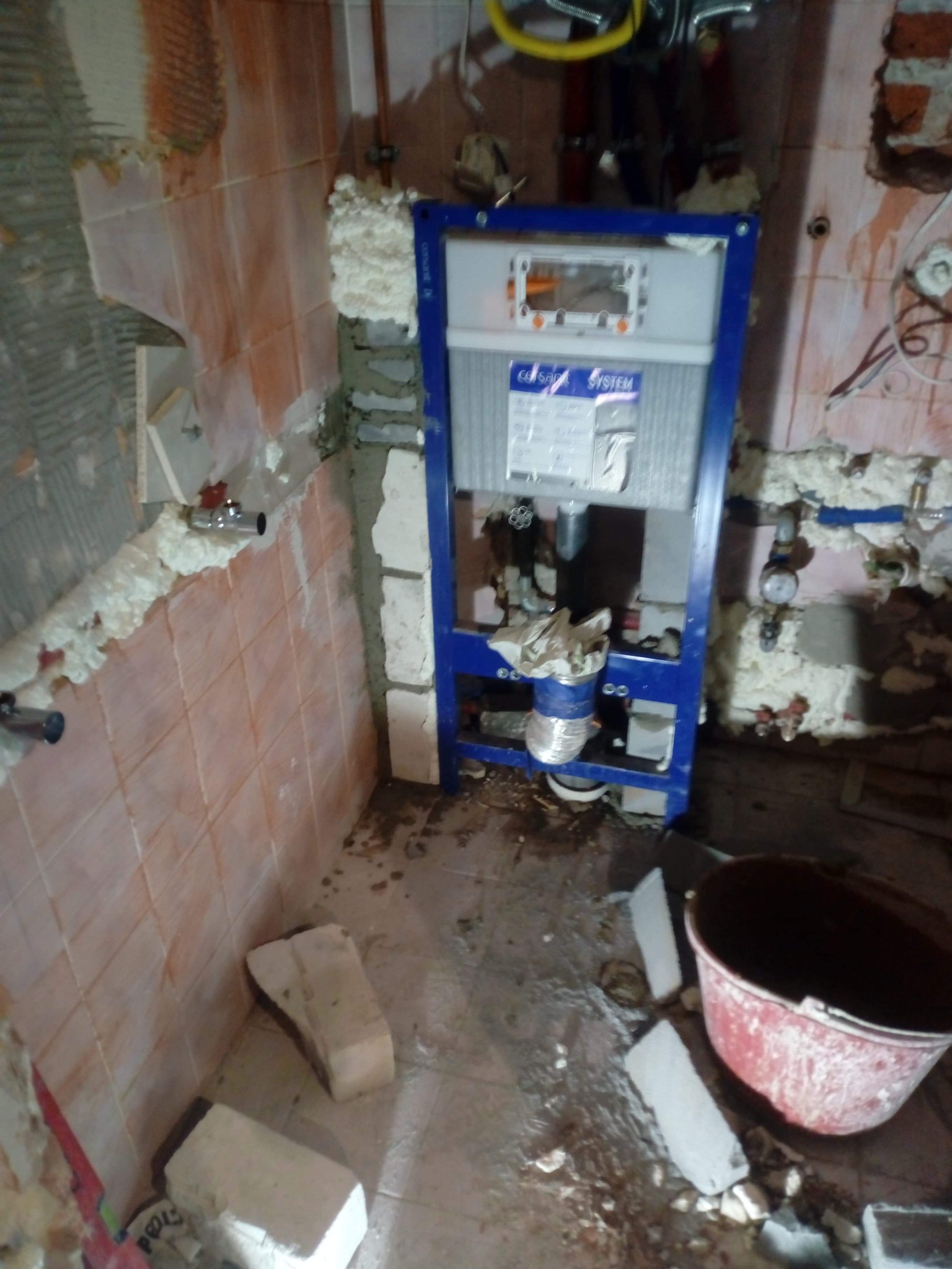 Montaż stelaża podtynkowego WC w trakcie remontu łazienki, widoczne fragmenty starej glazury, pianka montażowa i wiadro z zaprawą.