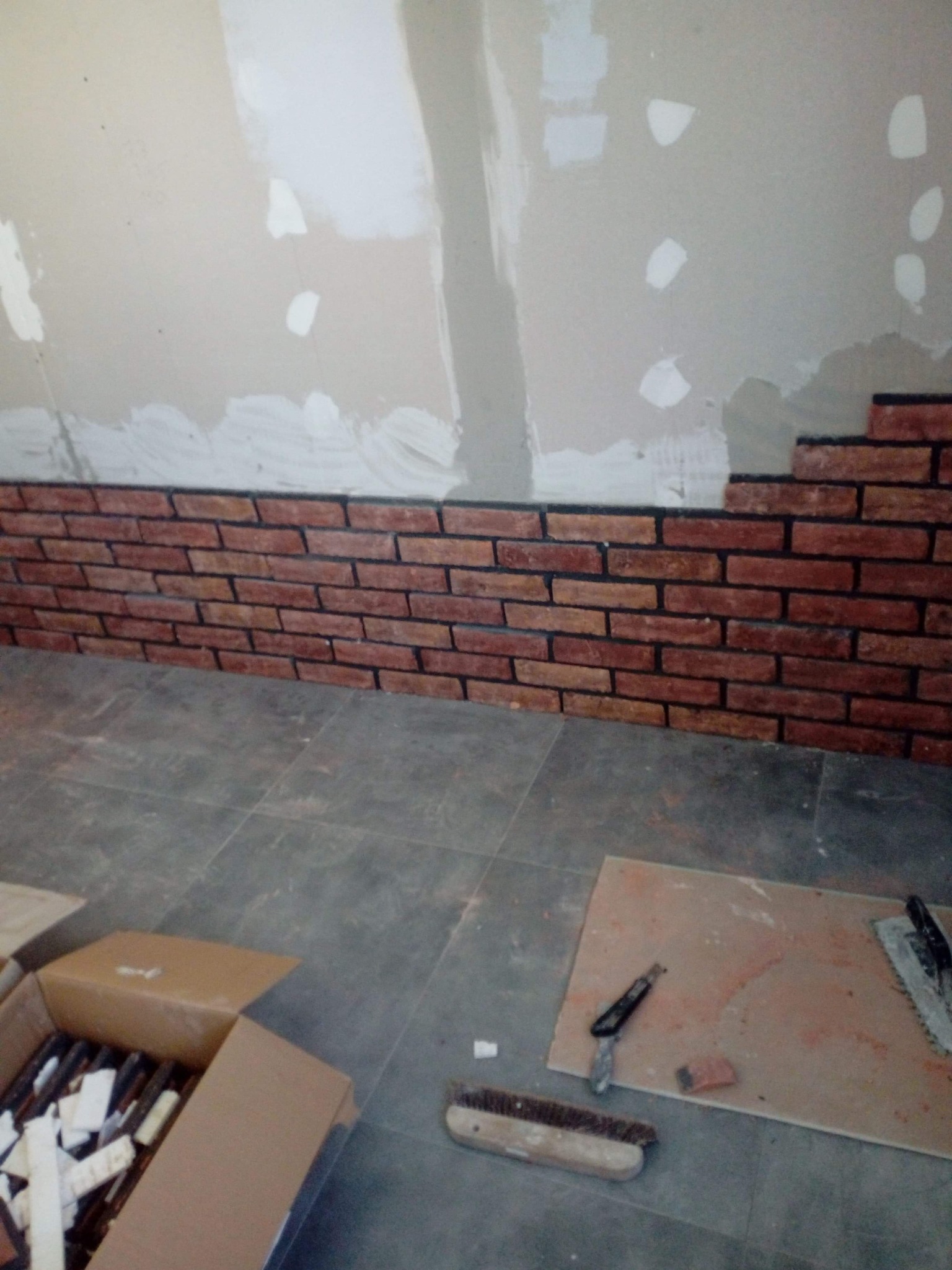 Układanie dekoracyjnej cegły na ścianie, widoczne narzędzia murarskie i karton z cegłami, ściana częściowo przygotowana, podłoga wyłożona płytkami.