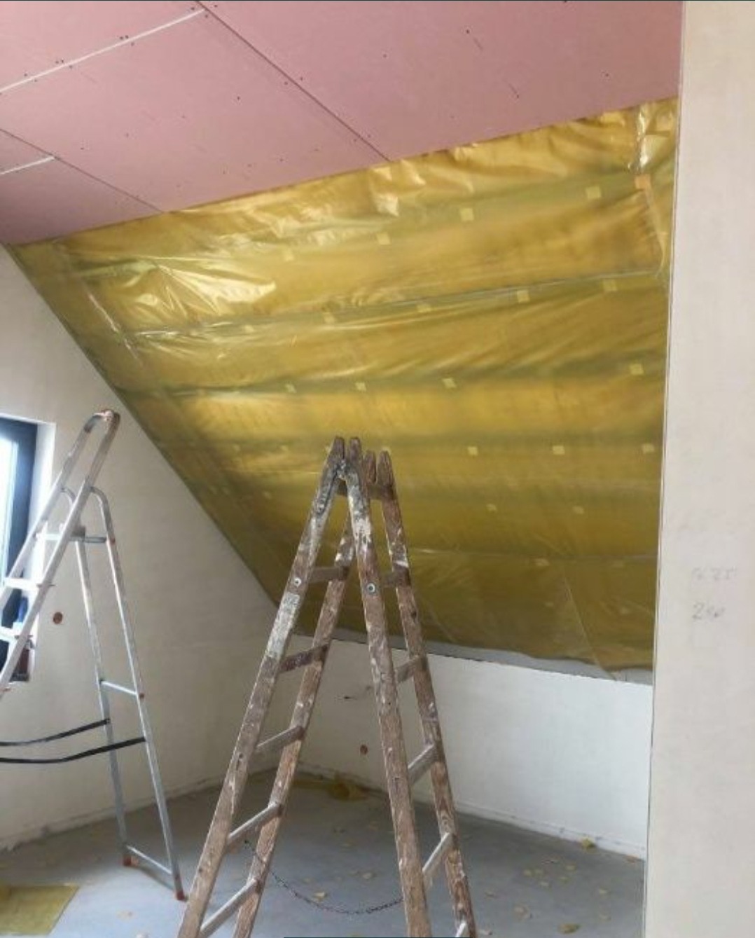 Poddasze w trakcie ocieplania, widoczna żółta folia zabezpieczająca izolację, dwie drabiny opierają się o ścianę.