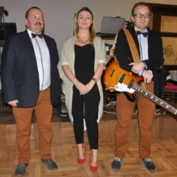 Trzyosobowy zespół na scenie: mężczyzna z wąsami w granatowej marynarce i muszce, kobieta w czarnym kombinezonie i czerwonych szpilkach oraz mężczyzna w okularach z gitarą elektryczną, za nimi...