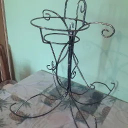 Metalowy, ręcznie robiony kwietnik z giętych prętów, stojący na stole z obrusem w ananasy, na tle zielonej ściany.