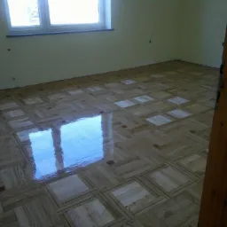 Świeżo polakierowana, błyszcząca podłoga parkietowa w geometryczny wzór, odbijająca światło okna, widok z perspektywy narożnika pokoju, fragment drewnianej framugi drzwi po prawej.