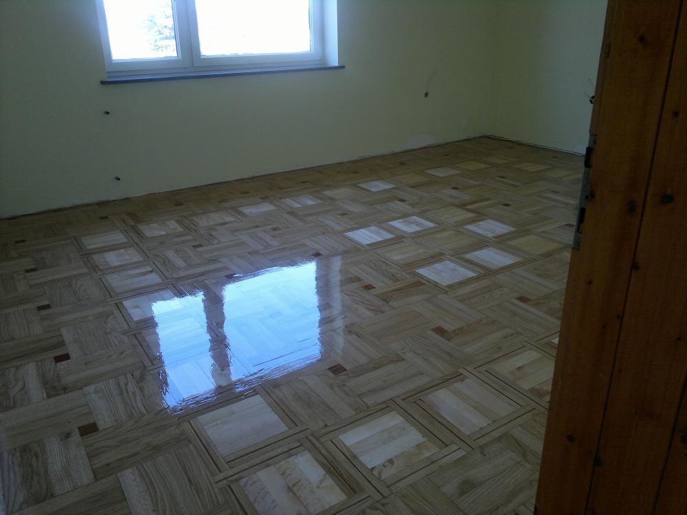 Świeżo polakierowana, błyszcząca podłoga parkietowa w geometryczny wzór, odbijająca światło okna, widok z perspektywy narożnika pokoju, fragment drewnianej framugi drzwi po prawej.