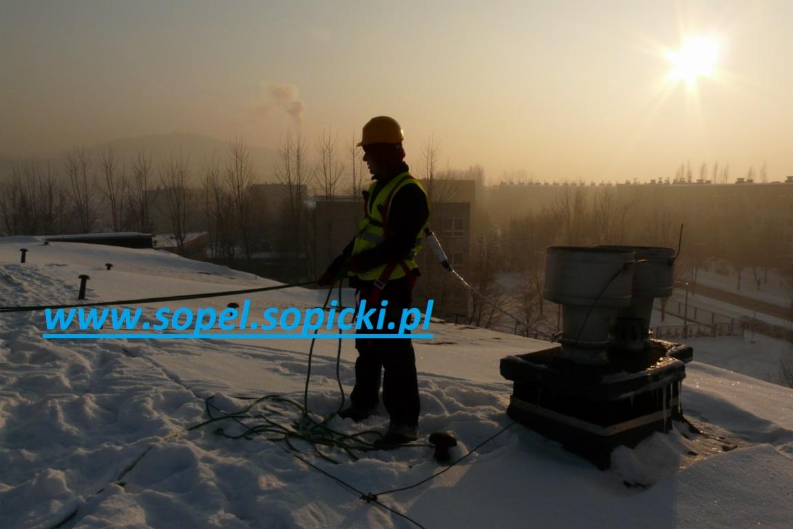 Pracownik w kasku i kamizelce odblaskowej, zabezpieczony linami, stoi na zasypanym śniegiem dachu budynku i przygotowuje się do pracy, prawdopodobnie usuwania śniegu lub sopli, z widocznym kominem...