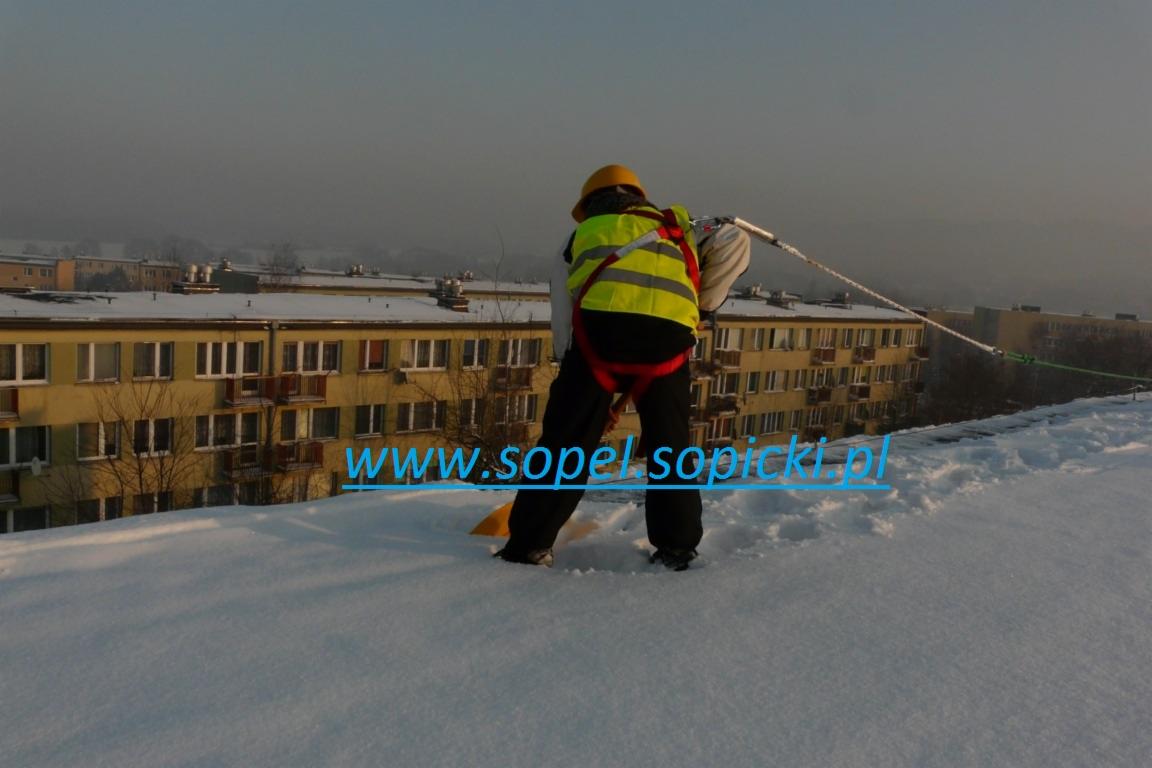 Pracownik w żółtym kasku i kamizelce odblaskowej, zabezpieczony linami, odśnieża płaski dach bloku mieszkalnego przy użyciu żółtej łopaty. Na dachu leży gruba warstwa śniegu.