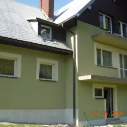 Dwukondygnacyjny dom z zieloną elewacją i białymi obramowaniami okien, widoczny z boku, z dachem z blachy falistej i brązowymi rynnami. Na elewacji widoczny datownik 07.05.2008.