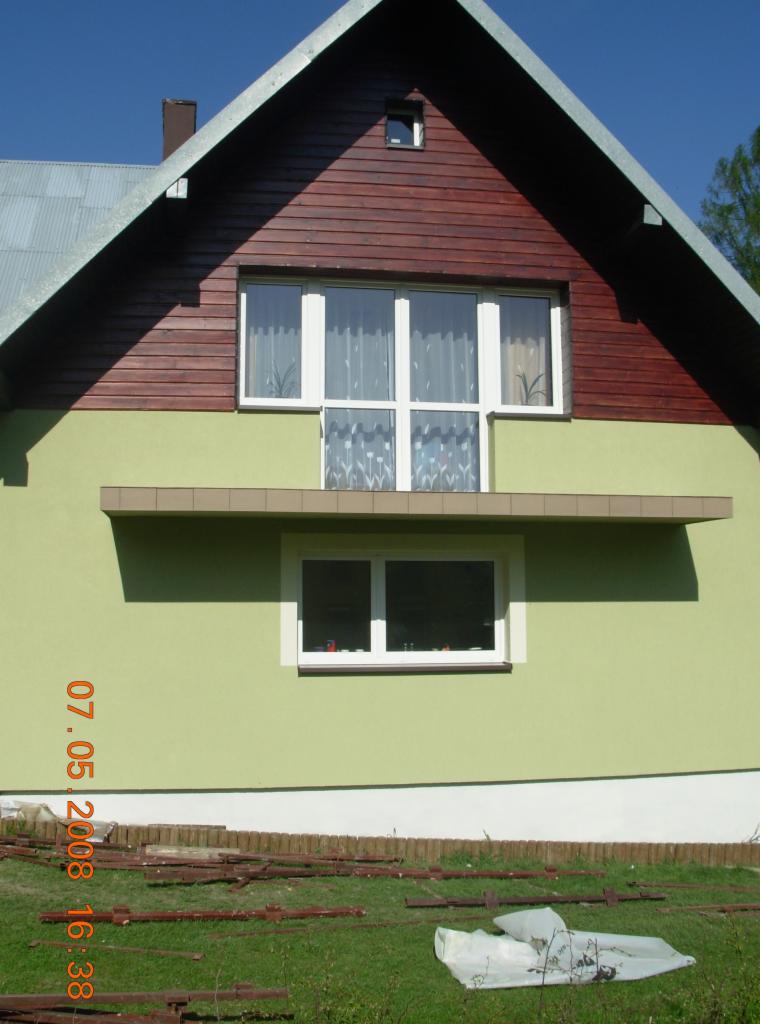 Fragment fasady domu z oknami, zieloną elewacją i drewnianym szczytem, widoczne elementy docieplenia na trawniku.