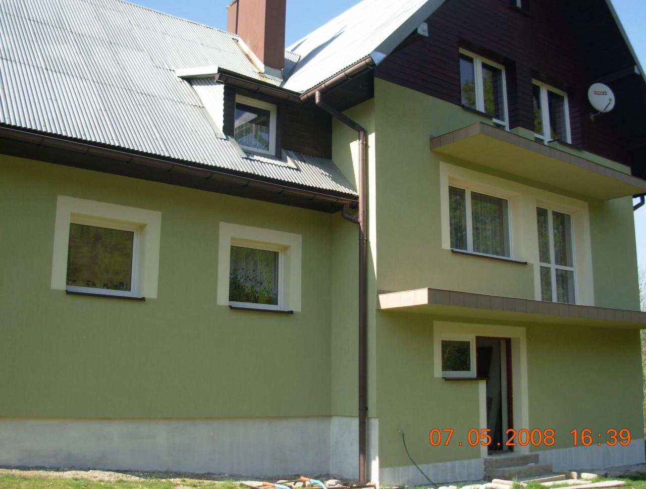 Dwukondygnacyjny dom z zieloną elewacją i białymi obramowaniami okien, widoczny z boku, z dachem z blachy falistej i brązowymi rynnami. Na elewacji widoczny datownik 07.05.2008.