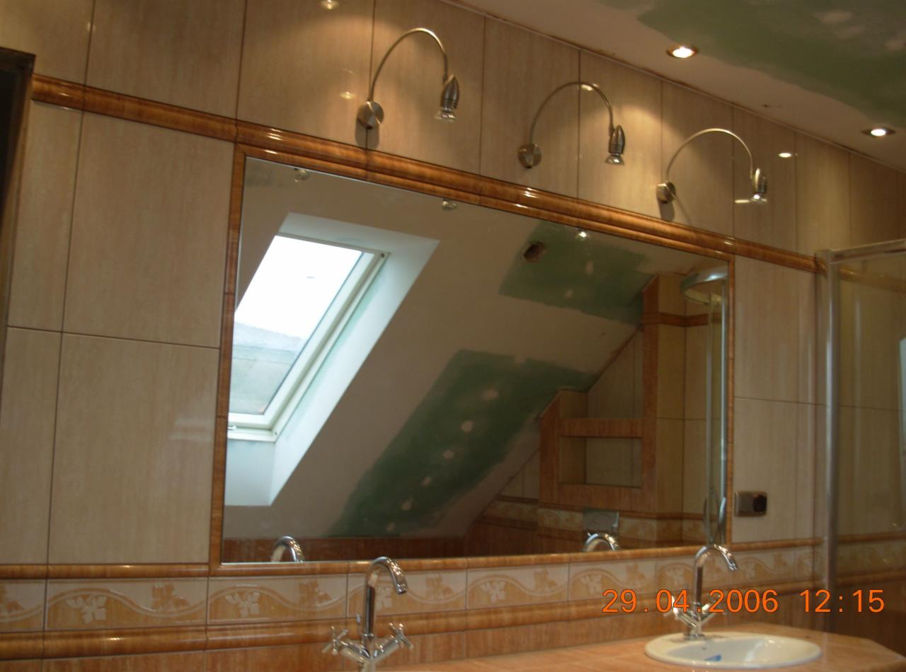 Łazienka z beżowymi płytkami, dużym lustrem z drewnianą ramą, trzema lampami nad lustrem i oknem dachowym widocznym w odbiciu, w trakcie prac wykończeniowych, widoczne ślady szpachli na ścianie.