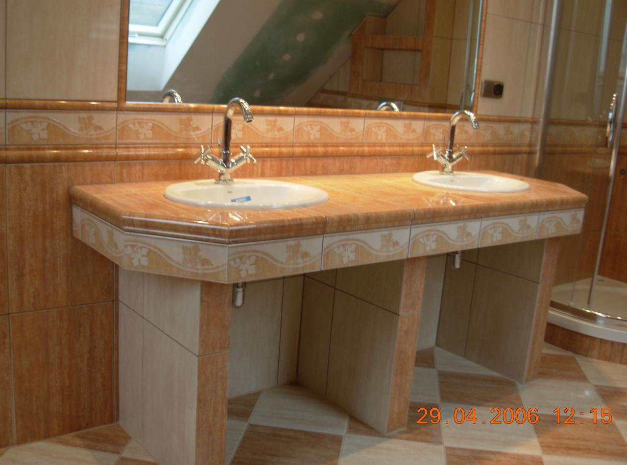 Umywalka podwójna z bateriami w łazience wykończonej płytkami w odcieniach beżu i brązu, widoczne lustro i kabina prysznicowa.