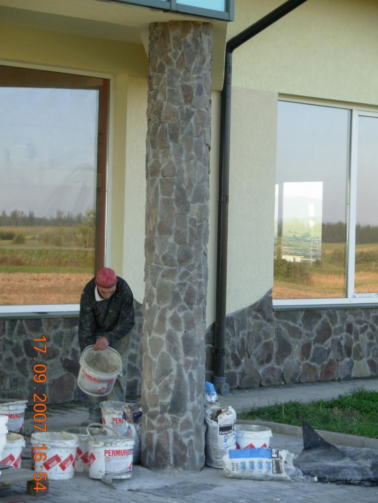 Pracownik w czerwonej czapce przygotowuje zaprawę przy kolumnie obłożonej kamieniem elewacyjnym, widoczne worki i wiadra z materiałami budowlanymi, fragment elewacji budynku z oknami.