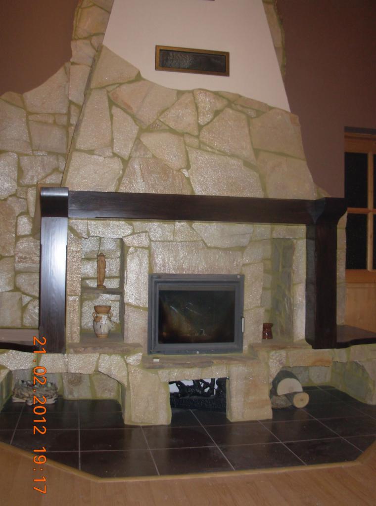 Kominek obudowany kamieniem z drewnianą belką, z zamkniętym paleniskiem i półkami na dekoracje, na podłodze ciemne płytki.