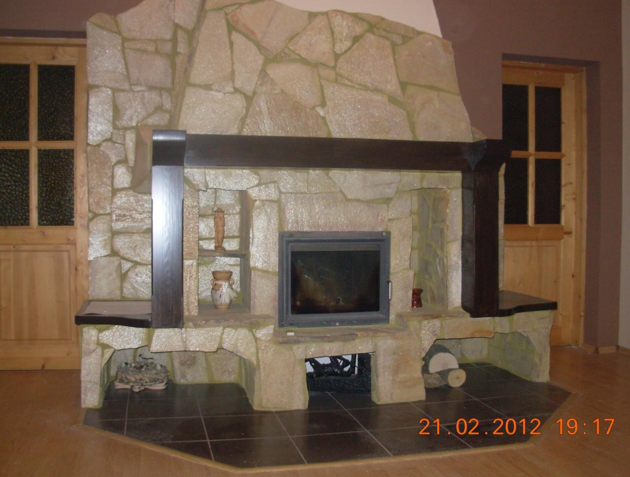 Kamienny kominek z drewnianą belką i półkami, obok drewniane drzwi z szybami, data na podłodze