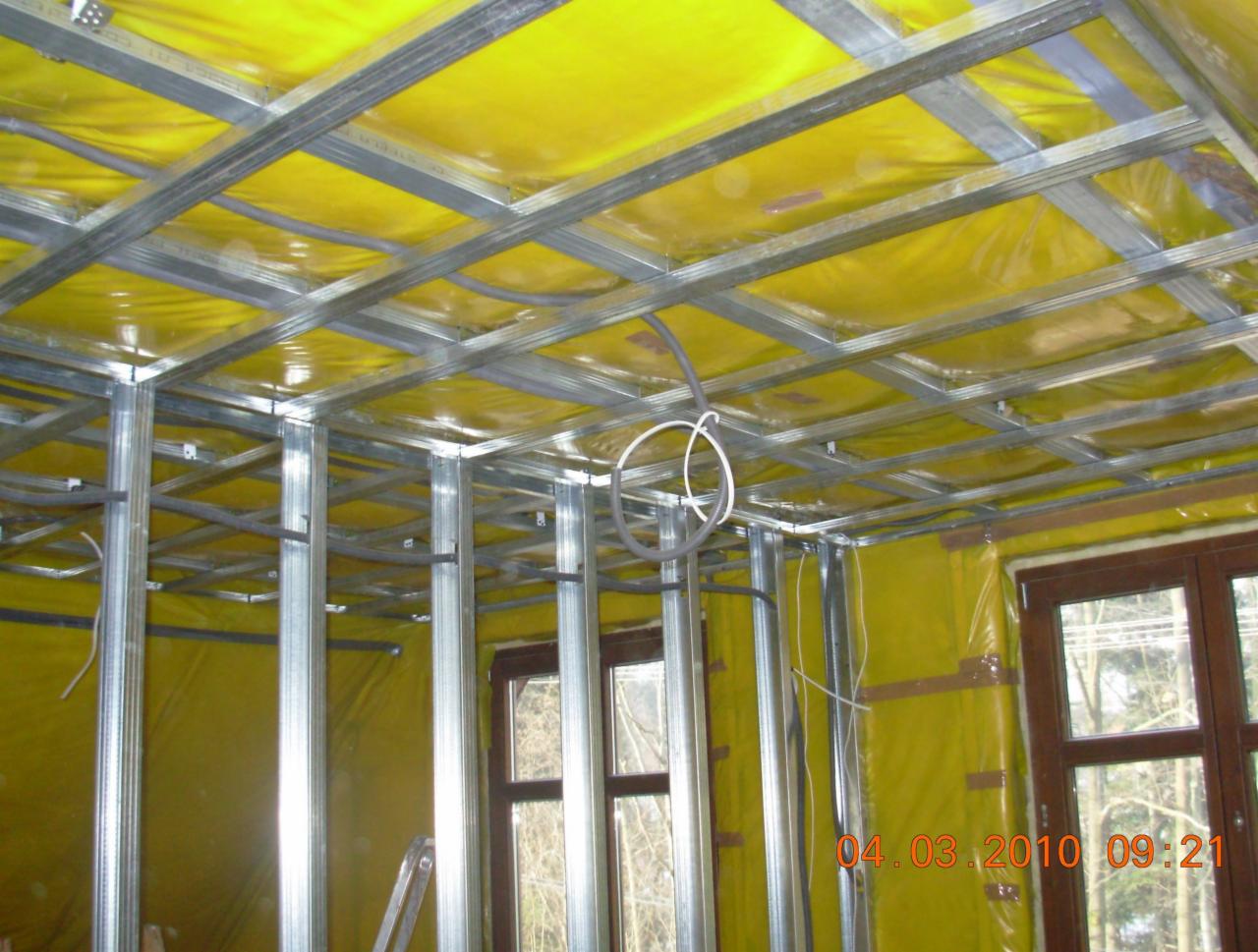 Montaż metalowej konstrukcji sufitu podwieszanego z widoczną izolacją w kolorze żółtym, instalacja elektryczna w pętli i okna z ciemnego drewna