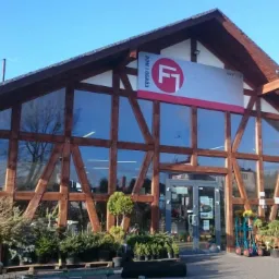 Fasada budynku z drewnianą konstrukcją i dużymi oknami, przed wejściem ekspozycja roślin w donicach.