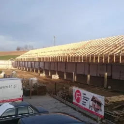 Budowa hali z drewnianą więźbą dachową w słoneczny dzień. Widoczny dźwig, samochody firmowe i materiały budowlane. Prace w toku.