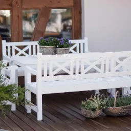 Biały zestaw mebli ogrodowych z drewna na tarasie, dekorowany kwiatami w koszykach, z charakterystycznym wzorem krzyży na oparciach. Ustawiony na brązowej podłodze tarasu.