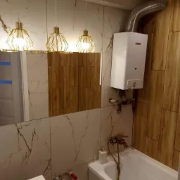 Nowoczesna łazienka po remoncie z białymi i drewnopodobnymi płytkami, złotymi lampami wiszącymi i białym bojlerem nad wanną.