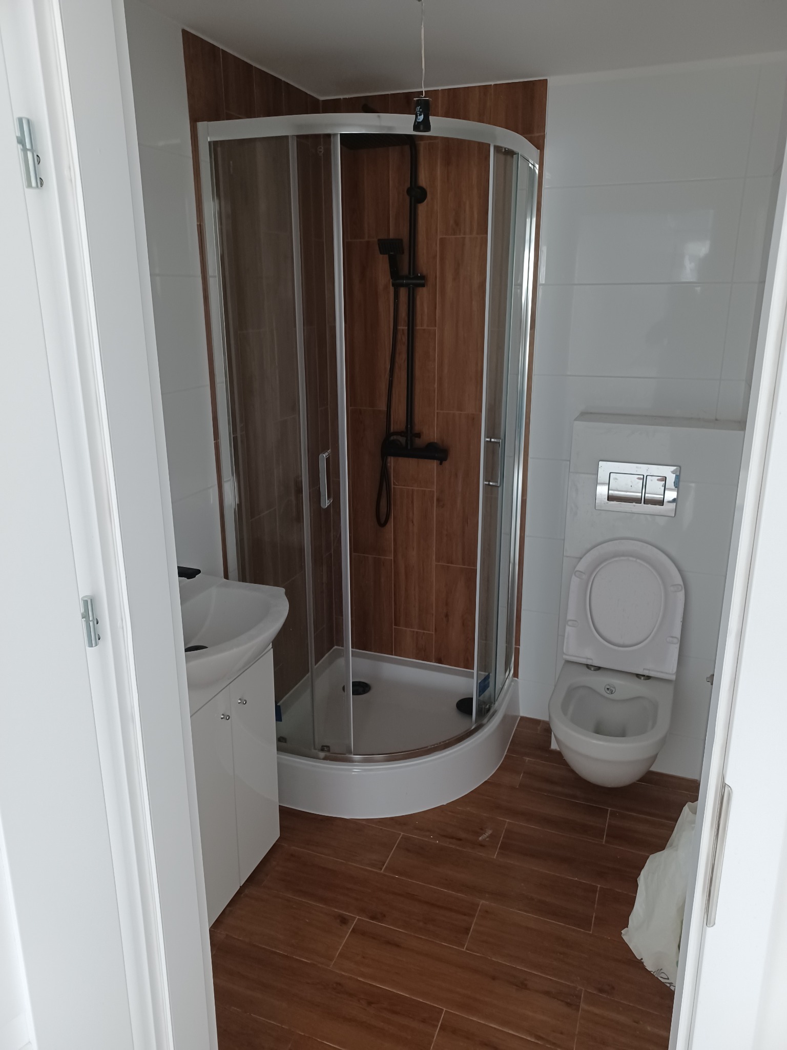 Nowoczesna łazienka z brązowymi płytkami podłogowymi imitującymi drewno, kabiną prysznicową z czarną armaturą, umywalką z białą szafką i podwieszaną toaletą.