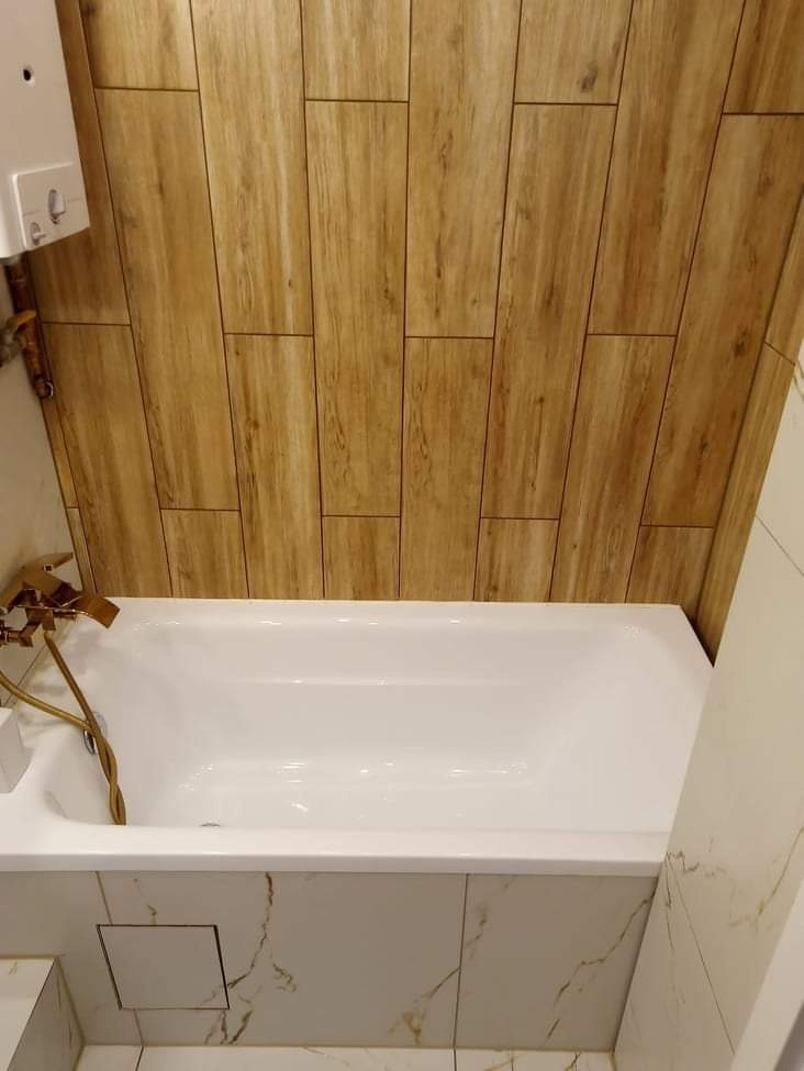 Biała wanna obudowana marmurowymi płytkami, z baterią w kolorze szczotkowanego złota, na ścianie okładzina z płytek imitujących drewno, widoczny biały bojler.