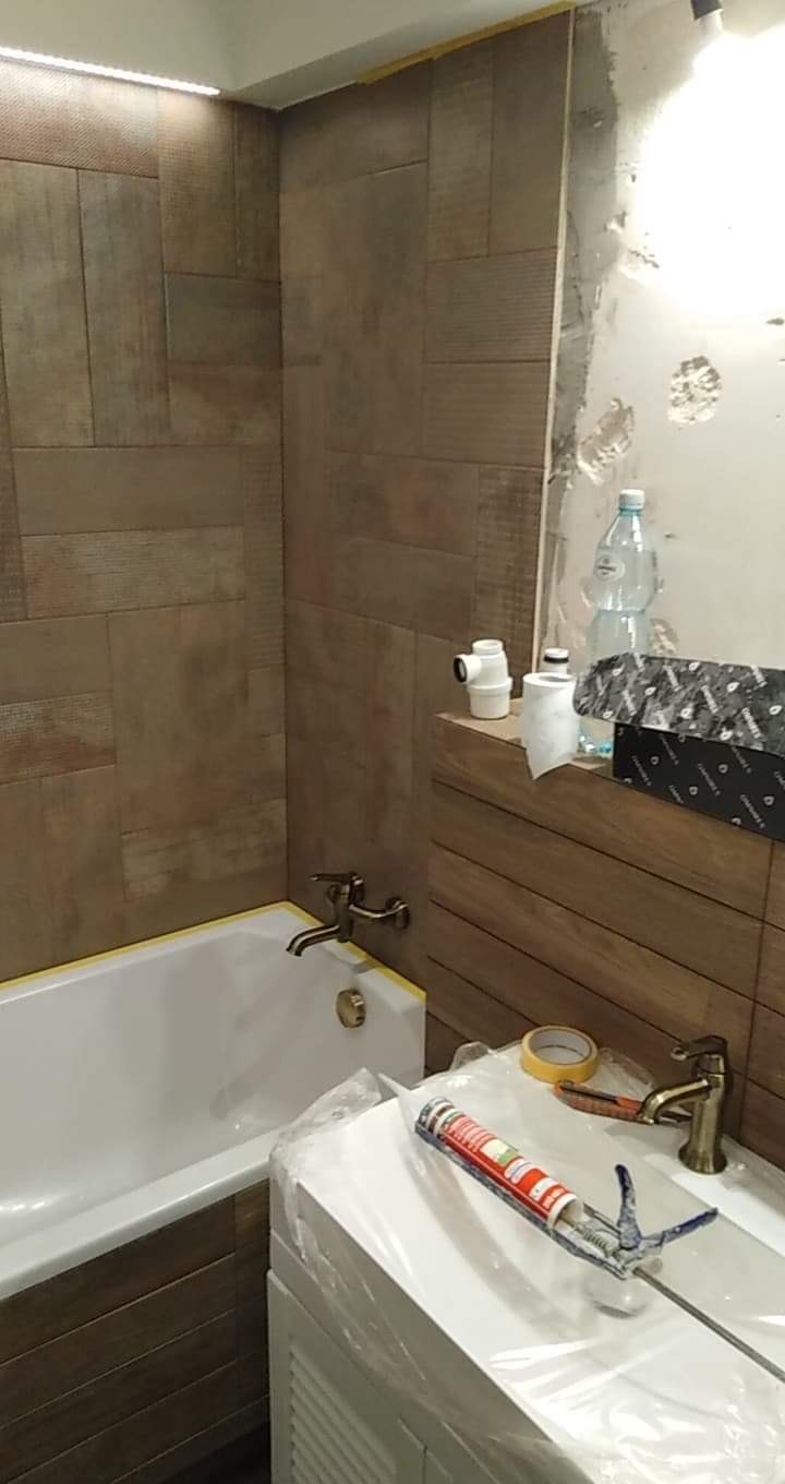 Łazienka w trakcie prac wykończeniowych: brązowe płytki imitujące drewno na ścianach, biała wanna z żółtą taśmą zabezpieczającą, umywalka owinięta folią ochronną, pistolet do silikonu na umywalce...