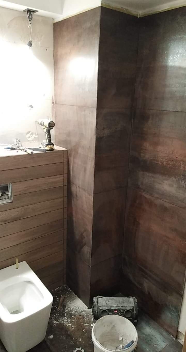 Wnętrze łazienki w trakcie prac wykończeniowych: częściowo ułożone płytki imitujące drewno na ścianach, wisząca toaleta, narzędzia budowlane i wiadro z zaprawą na podłodze.