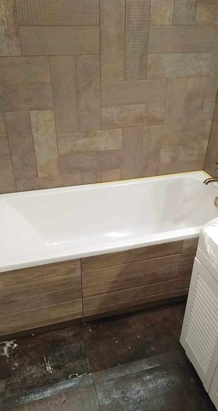 Biała wanna obudowana panelami imitującymi drewno, z brązowymi płytkami na ścianie ułożonymi w nieregularny wzór, widoczna pralka i zabrudzona podłoga w trakcie prac wykończeniowych.