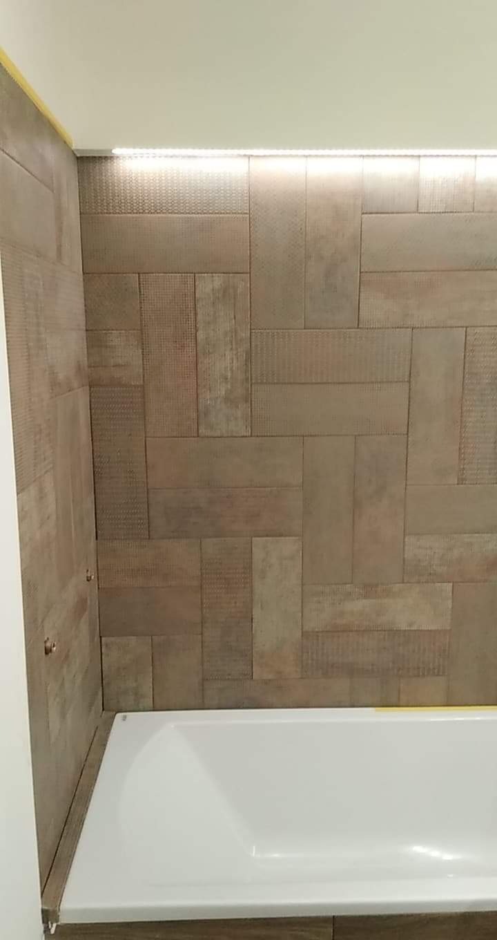 Wykończona łazienka z beżowymi płytkami o zróżnicowanej fakturze, ułożonymi w nieregularny wzór na ścianie nad białą wanną, podkreślona subtelnym oświetleniem LED podsufitowym.