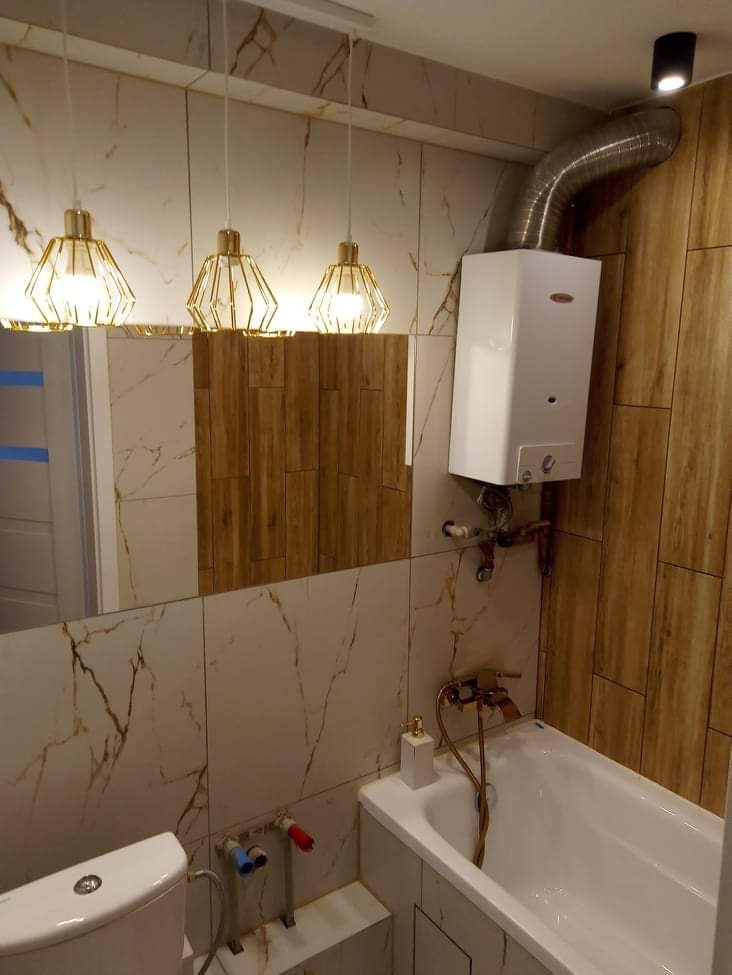 Nowoczesna łazienka po remoncie z białymi i drewnopodobnymi płytkami, złotymi lampami wiszącymi i białym bojlerem nad wanną.