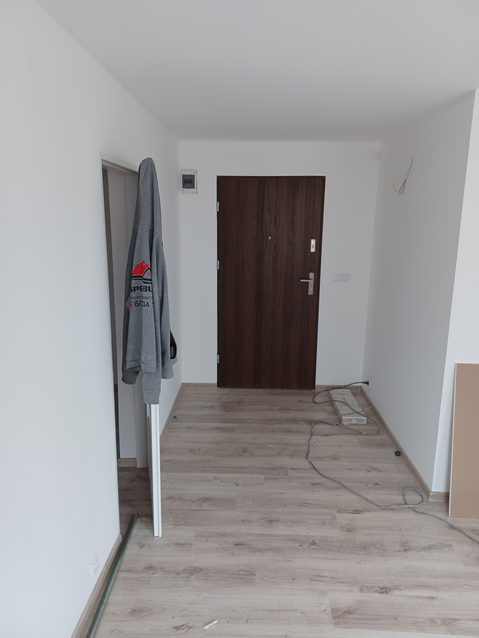 Nowe panele podłogowe w przedpokoju z brązowymi drzwiami wejściowymi, szara bluza z logo firmy remontowej wisi na framudze, widoczne listwy przypodłogowe i przewody elektryczne.