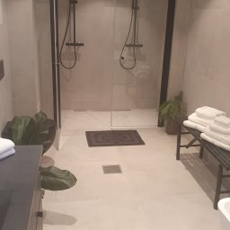 Minimalistyczna łazienka z dwoma kabinami prysznicowymi typu walk-in, beżowe płytki podłogowe i ścienne, czarne armatury, rośliny doniczkowe i sterta białych ręczników.