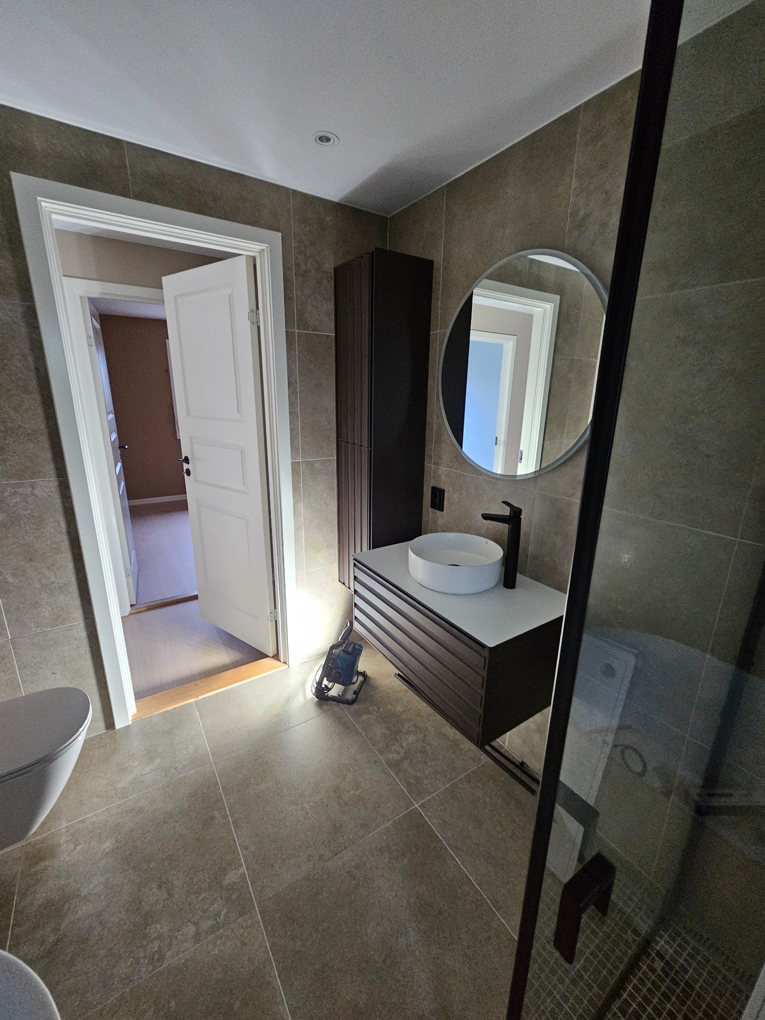 Nowoczesna łazienka z umywalką na szafce, kabiną prysznicową i otwartymi drzwiami. Widoczna płytka podłogowa i ścienna w odcieniach szarości. W tle widoczny pokój.