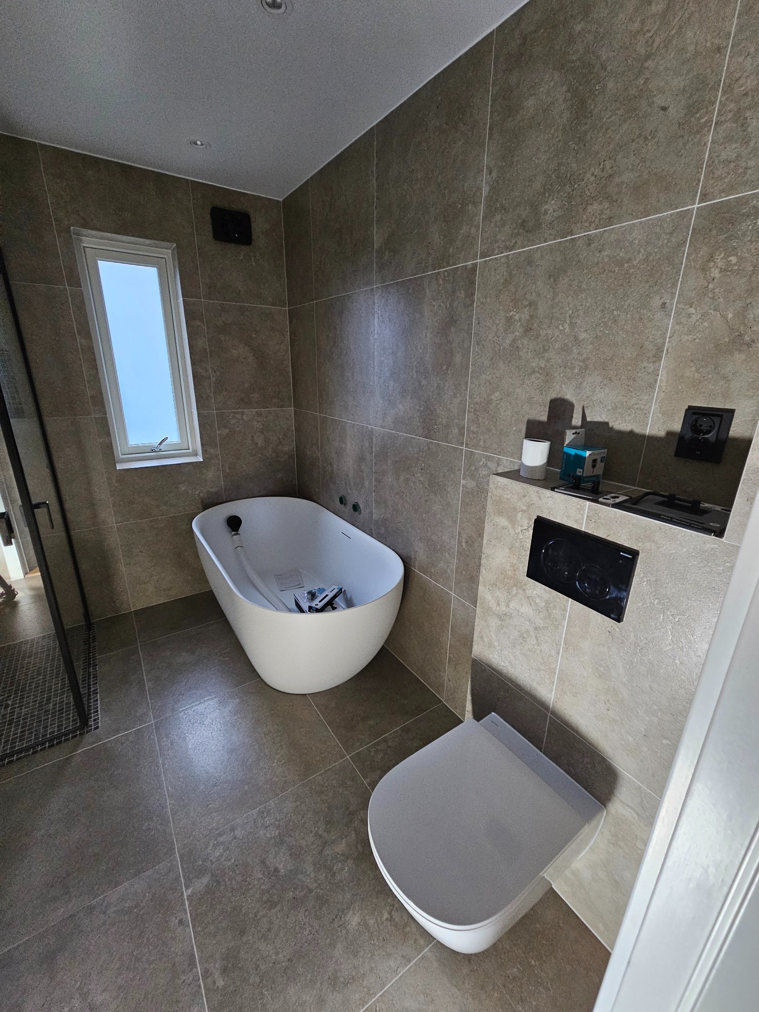 Nowoczesna łazienka z wanną wolnostojącą, toaletą i szarą glazurą. Widoczne elementy instalacji przed zabudową. Okno z widokiem na niebo.